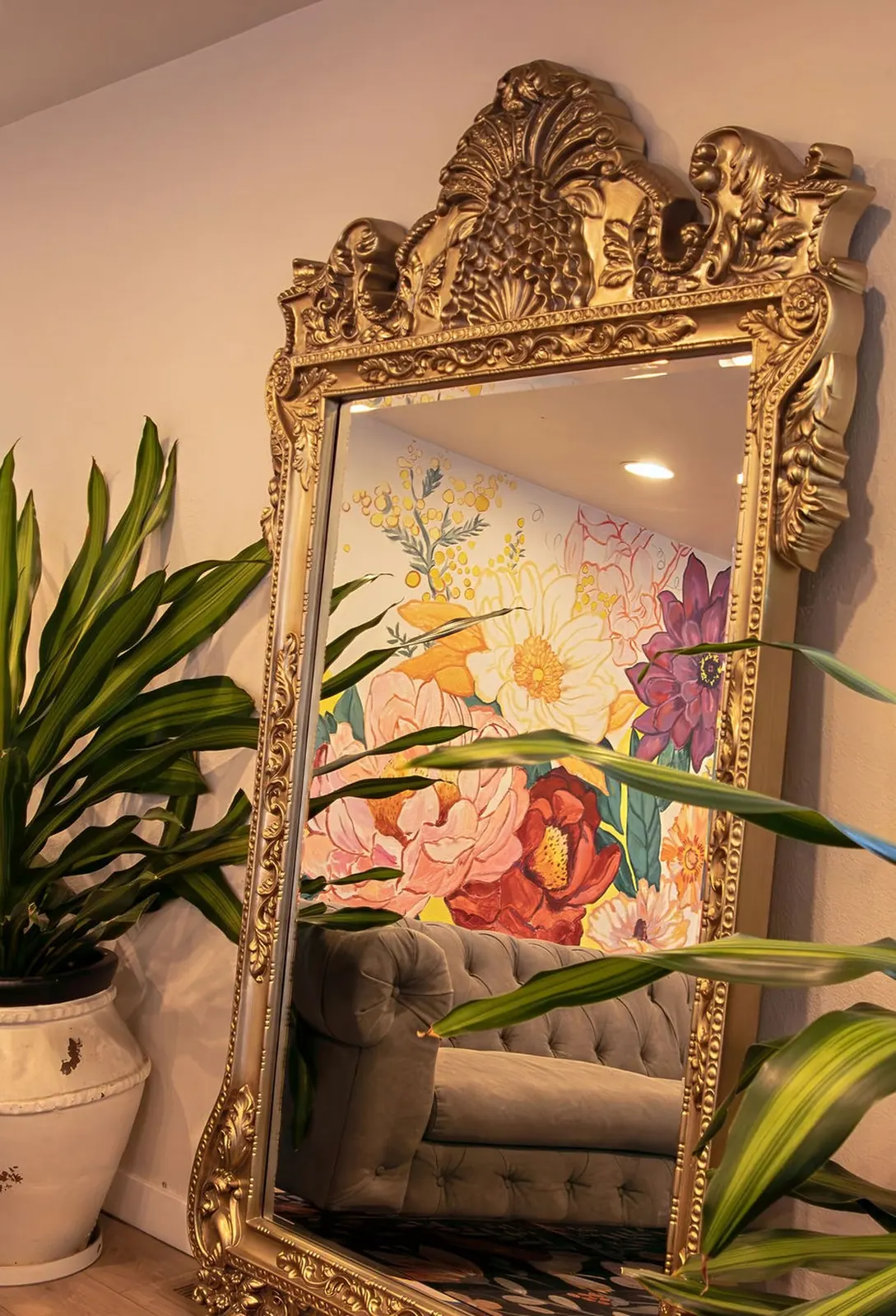 A gilded floor mirror at Wax &amp; Lash Berkeley reflecting the hand painted floral mural, surrounded by tall green houseplants